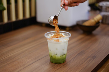 pouring corn flakes into a plastic glass of matcha tea and ice cubes for making iced matcha green...