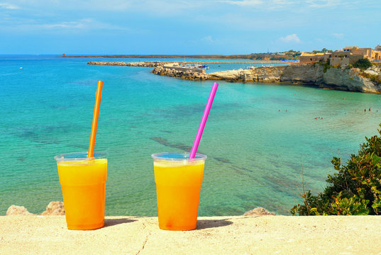 bright orange cold refreshing smoothie drink in a plastic cup on stone wall on beach of old coastal mediterranean town Terrasini with turquoise sea water and bright summer day,  Palermo, Sicily Italy