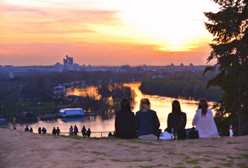 four young girls on Kalemegdan fortress park hill looking warm colorful sunset on Danube river  with horizon of New Belgrade in background in Belgrade, Serbia - obrazy, fototapety, plakaty