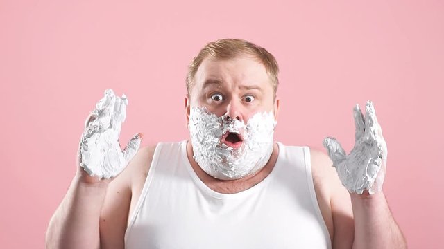 Upset man with gel on cheeks, has sad expression, sensitive skin, man going to shave his chin despite ofskin irritation, isolated over pink background, slow motion.