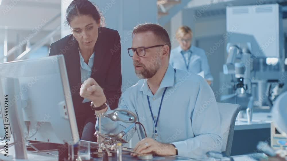 Poster female chief engineer talks with electronics specialist, explaining things, he works on computer and