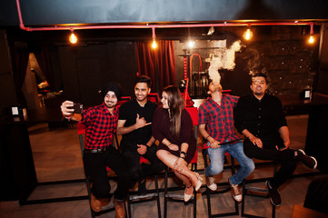 Group of indian friends sitting at lounge bar, having fun and rest, smoking hookah and looking on mobile phones to make selfie.