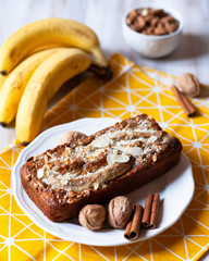 Delicious homemade banana cake. Served on a white plate and yellow napkin. Decorated with cinnamon and almond powder. Bananas and bowl with almonds on background. Light table. Morning
