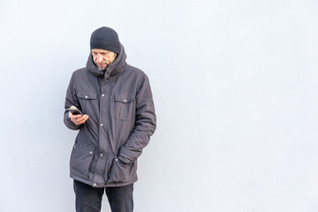 a man in a jacket and hat stands near a single-color wall and looks into the screen of a smartphone, copy space
