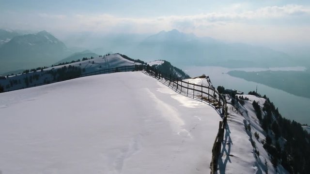 A drone movie over a mountain cliff with a stuning scenic view to a lake.
A fence is visible on the edge of the cliff. A fantastic movie on a winterday in the mountains.