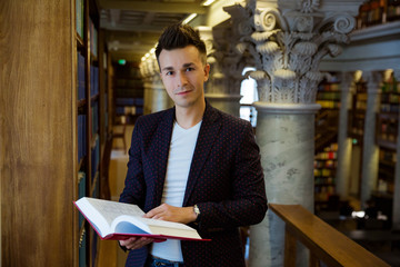 Young handsome man standing in traditional library at bookshelves, holding book. Beautiful interior of old library with pillars. Higher education. 
