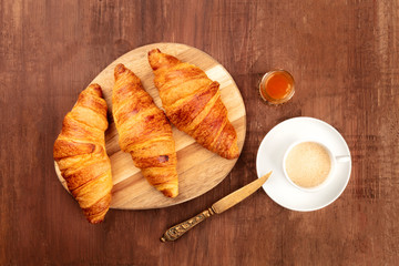 A photo of three croissants, shot from above on a dark rustic wooden background with coffee, jam, and a place for text