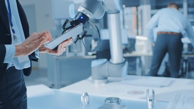 Stylish Female Industrial Robotics Engineer Uses Tablet Computer to Program and Manipulate Robot Arm for Lifting, Moving Objects. Technology Research Facility for Machine Learning and Cloud Computing