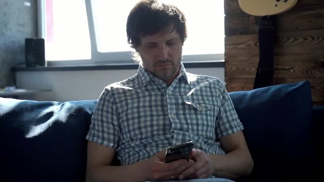 Slowmotion. A man sits on the couch at home and prints a message on the phone connected to the Internet via Wi-Fi. Front view