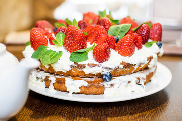 Strawberry cake in cream and mint leaves on the table in a plate, on the background of a teapot with tea, in light colors, close-up with a blurred background.