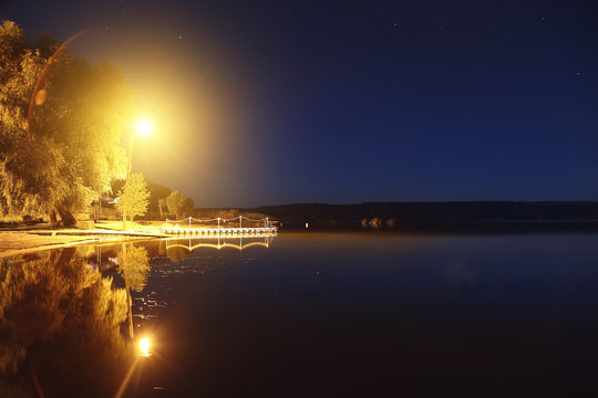 Lake or river sand shore with trees and glowing lantern over pantoon pier and dark blue starry sky on background. Tranquil nature night landscape