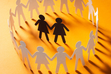 Paper figure of female couple surrounded by circle of paper people holding hands on yellow surface. Bulling, minorities, conflict concept.