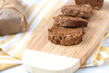 Homemade Zucchini Bread Sliced on Bamboo Cutting Board; Second Loaf Wrapped in Parchment Paper; Yellow and White Dish Towel on White Countertop