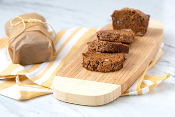 Homemade Zucchini Bread Sliced on Bamboo Cutting Board; Second Loaf Wrapped in Parchment Paper; Yellow and White Dish Towel on White Countertop