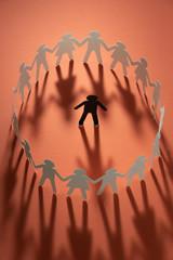 Paper human figure surrounded by circle of paper people holding hands on red surface. Bulling,...