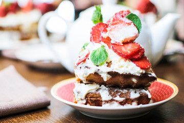 A piece of strawberry cake in cream and mint leaves on a table in a plate, in front of a teapot with tea, in light colors, close-up with a blurred background.