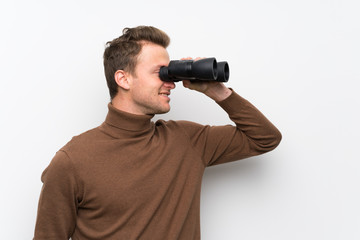 Blonde man over isolated white wall and looking in the distance with binoculars