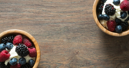 Delicious sweet dessert made from berry mix (raspberries, blueberries and blackberries) with whipped cream in a wooden bowl on a wooden background. Close up.