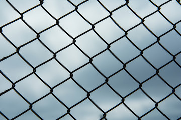 Metal black fence-mesh netting. Gloomy cloudy gray sky. The background image of the fence.