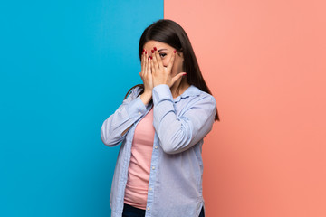 Young woman over pink and blue wall covering eyes by hands and looking through the fingers