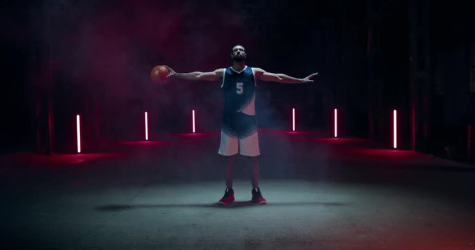 CINEMAGRAPH - SEAMLESS LOOP. African American professional basketball player posing with a ball against dark background in a large abandoned warehouse. 4K UHD