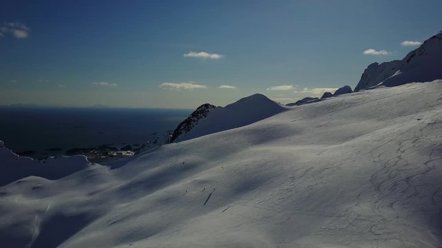 drone from far,wide shot of snowkiters in snowy hill on a sunny day at lofoten norway .huge snow covered mountains.blue sky with clouds.icy ocean at distance.travel getaway on norway. 4k 30fps