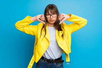 Young woman with yellow jacket on blue background showing thumb down with both hands