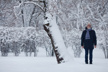 Full length portrait of handome mature man in trendy winter clothing and fashionable glasses standing over snow park background. Outdoor shot