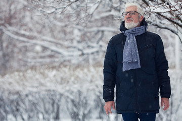 Active grandpa concept. Portrait of handome mature man in trendy winter clothing and fashionable glasses standing over snow park background. Copy-space.  Outdoor shot