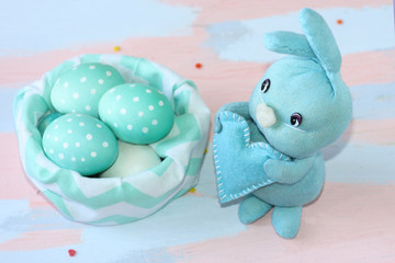Easter light blue rabbit sewn by hands near a cloth basket with blue-white eggs . Handwork. Greeting card for happy easter.