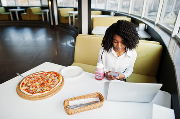 Сheerful business african american lady with afro hair, wear white blouse sitting at table, work with laptop in cafe, and drink pink milkshake cocktail, eat pizza, look on mobile phone.