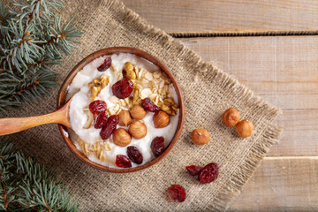 Wooden bowl with yogurt with rolled oats, nuts and dried cranberries on burlap on wooden table.