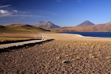 View on track along deep blue lake at Altiplanic Laguna (Lagoon) Miscanti in Atacama desert with partly snow capped cones of volcano Meniques in the background - Chile