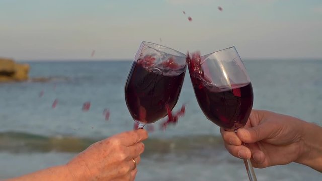 Zeitlupenaufnahme mit zwei Gl&auml;sern Wein, die ansto&szlig;en. Der Rotwein schwappt &uuml;ber mit dem Strand und dem blauen Meer im Hintergrund.