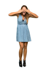 A full-length shot of a Blonde woman with jean dress covering eyes by hands. Surprised to see what is ahead over isolated white background