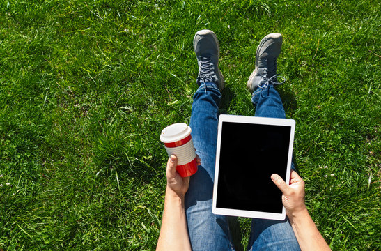 Top view of man in casual outfit using laptop while sitting on green grass with digital tablet and cup of tea/coffee. Concept of freelance job or study, copy space.