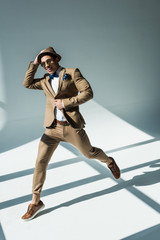 stylish mixed race man in suit smiling and jumping on grey with sunlight