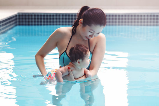 Mixed Race Asian Mother Traning Her Newborn Baby To Float In Swimming Pool. Baby Diving In Water. Healthy Active Lifestyle. Family Activity And Early Development Concept