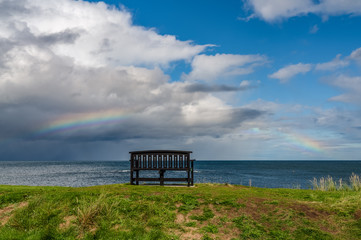 A bench with a Rainbow over the North Sea coast, seen in Benthall, Northumberland, England, UK