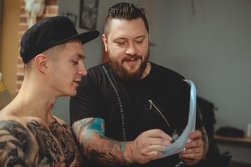 Bearded professional tattoo artist showing his customer tattoo sketches at his studio. Young...