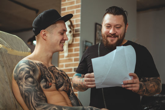 Cheerful bearded tattoo artist smiling joyfully talking to his client. Young tattooed man discussing sketches with his tattooist before getting inked. Professional tattoo artist working with a client