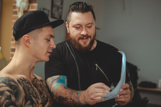 Bearded professional tattoo artist showing his customer tattoo sketches at his studio. Young tattooed man preparing to get a new body tattoo. Tattooist talking to his client before tattooing