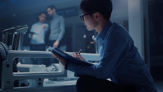 Automobile Designer Sitting Beside Hybrid Electric Car Chassis Platform, Writes Estimations and Sketches in the Notebook. Facility with Vehicle Frame with Suspension, Wheels, Engine and Battery