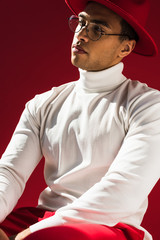 confident stylish mixed race man in hat and glasses posing isolated on red