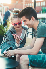 Teenage girl and boy,  having fun using smartphones sitting in center of town