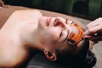 Close up portrait of a young caucasian woman with acne having anti acne mask with chocolate by a cosmetologist in a wellness spa.