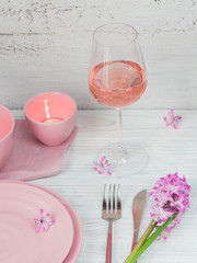 Pink rustic table setting with purple hyacinth flowers, linen napkin, candles and glass of rose wine on white wooden table