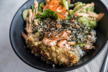 Poke Bowl mit flambierten Lachs, Tempura Shrimp, Guacamole, Masago Kaviar, Salat, Nori Algen und Sesam auf Reis in schwarz Porzellan Schale
