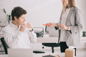 cropped view of blonde woman holding pills and glass of water near coworker with tissue