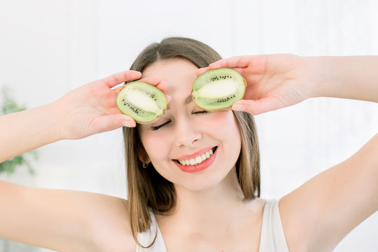 Cheerful beautiful young woman holding two cut kiwis on her eyes for fun joke , indoors studio shoot. Beauty and skin care concept